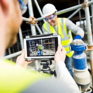 Travailleur en tenue de sécurité sur un chantier de construction manipule du matériel tandis qu'un collègue visionne sur une tablette les images capturées avec la solution 360SkillVue dans le cadre du processus d'habilitation.